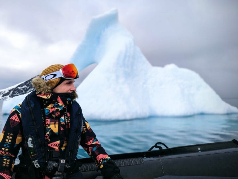 Man in winter gear rides on a zodiac boat in Antarctica with an iceberg in behind him